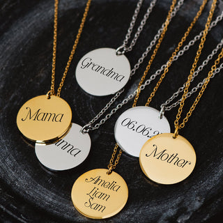 Collection of gold and silver personalized disc necklaces engraved with names and titles like ‘Mama,’ ‘Grandma,’ and ‘Mother,’ displayed on a dark surface