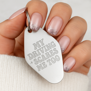 Close-up of a hand holding a white keychain that reads “My driving scares me too,” a humorous gift idea for her, with manicured nails and soft neutral tones
