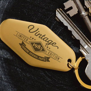 A close-up of a gold keychain tag attached to a set of keys, featuring the text “Vintage 1985 Limited Edition Genuine Original Parts” in a stylized design on a dark textured background