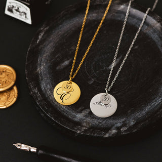 Gold personalizsed disc necklace with engraved script initial with charm, displayed on a dark textured surface.