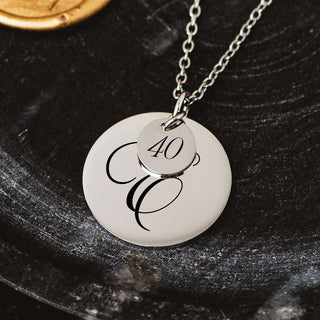 Silver personalised disc necklace with engraved script initial “E” and small “40” charm, styled on a dark textured surface.
