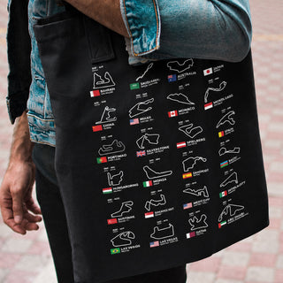 A person wearing a denim jacket and black pants carries a black tote bag printed with a grid of race track maps and small country flags.