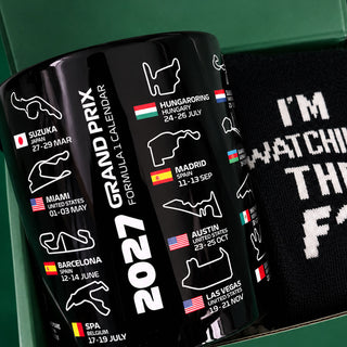 A black cylindrical mug printed with the 2027 Grand Prix calendar, shown alongside a black knit sock with white text inside a green gift box.