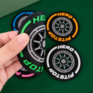 A hand holds a black tire-shaped accessory with green and white lettering, while three similar circular tire-shaped accessories rest on a green surface. The accessories feature the words HERO and PITSTOP in contrasting colors.