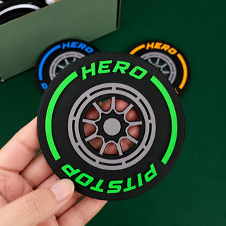 A hand holds a black circular pit stop tire-shaped accessory with bright green 'HERO' and 'PITSTOP' text and a gray wheel design. Two more matching accessories in blue and orange are blurred in the background on a green surface.