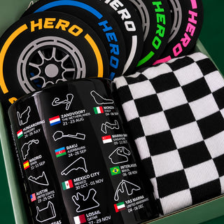 A close-up of a gift box containing a set of black crew socks with a black and white checkered pattern, alongside several black tire-shaped coasters featuring colorful 'HERO' lettering and race track names.
