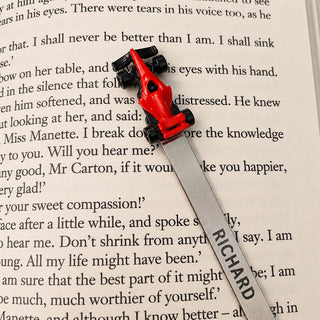 A silver metal bookmark with a red zinc alloy F1 car attached to a long silver metal bookmark printed with the name 'RICHARD', resting on an open book page.