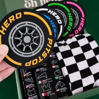 A close-up of a gift box containing a black and white checkered sock, a stack of circular coasters with colorful HERO branding, and a black track mat with race track illustrations. A hand is holding one of the coasters.