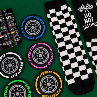 A flat-lay product photo showing a pair of black and white checkered crew socks alongside several circular tire-shaped coasters and a black can printed with racing graphics. The socks feature a black and white checkerboard pattern, while the coasters are black with colorful tire rims in blue, pink, yellow, green, and white, and the can displays various race flags and track names.