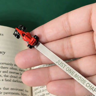 A hand holding a silver metal bookmark shaped like a miniature F1  car on a book page, with the bookmark printed with the phrase 'JUST ONE MORE CHAPTER'.