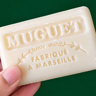 A hand holding a cream-colored bar of Muguet Fabriqué à Marseille soap against a solid dark green background.