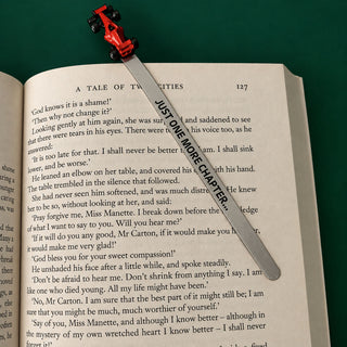 A silver metal bookmark with a red F1 car and a black printed text overlay reading 'JUST ONE MORE CHAPTER', shown resting on an open book page against a dark green background.