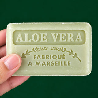 A hand holding a rectangular bar of pale green soap with embossed text reading 'ALOE VERA' and 'FABRIQUÉ A MARSEILLE' against a dark green background.