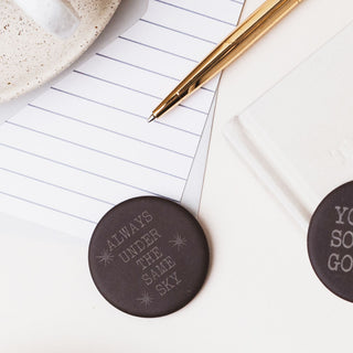 Two round dark brown coasters with engraved text, displayed on a white desk alongside a gold pen, lined paper, and a ceramic mug.