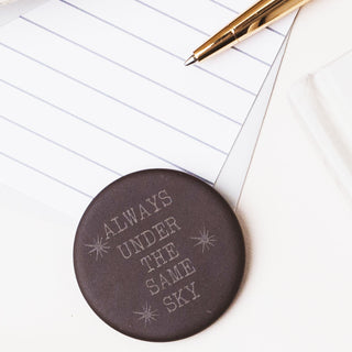 A round dark brown coaster with the phrase 'ALWAYS UNDER THE SAME SKY' and small starburst designs, styled on a white surface next to lined paper and a gold pen.