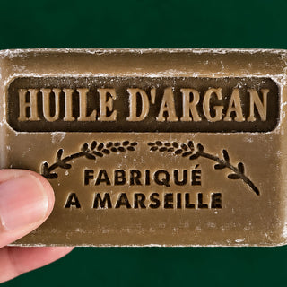 A close-up of a hand holding a rectangular bar of argan oil soap against a solid dark green background. The soap is a warm brownish-gold color with embossed French text reading 'HUILE D'ARGAN' and 'FABRIQUÉ A MARSEILLE'.