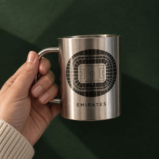 A hand holding a brushed stainless steel mug featuring a black stadium seating graphic and the word 'EMIRATES' on the front, set against a dark green background.