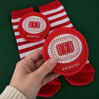 A hand holds a red circular Emirates stadium graphic coaster in front of red and white striped socks on a dark green background.