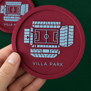 A hand holds a round burgundy coaster featuring a light blue stadium graphic and the text 'VILLA PARK'. Another coaster is visible in the background on a dark green surface.