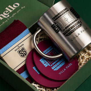 A gift set in a green box featuring a stainless steel mug with a black soccer field graphic and 'VILLA PARK' text, a maroon and light blue striped knit sock with a branded paper band, and two matching maroon coasters with blue graphics and 'VILLA PARK' text, arranged on shredded paper.