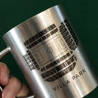 A close-up of a brushed stainless steel mug being held by a hand, featuring a black laser-engraved graphic of a soccer stadium and the text 'VILLA PARK'.