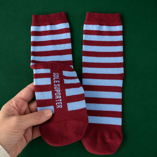 A pair of red and light blue striped crew socks, with one sock folded over to show the sole. The sole features the text 'SOLE SUPPORTER' in white.