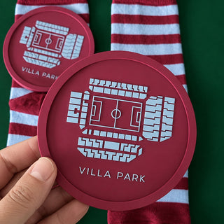 A hand holds a round burgundy coaster featuring a white stadium graphic and the text 'VILLA PARK'. In the background, claret and blue striped socks are laid out on a dark green surface, with another coaster partially visible.