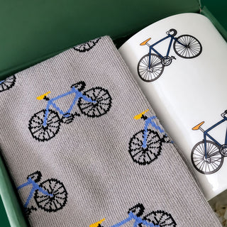 A close-up of a gray knit fabric with a repeating bicycle pattern in blue, black, yellow, and orange, placed next to a white ceramic mug with a matching bicycle print, inside a green box.