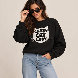 A woman wearing a black crewneck sweatshirt with a white graphic that reads 'Crazy Cat Lady' inside a cat head outline, paired with light blue jeans and black sunglasses, against a plain beige background.