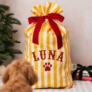 A yellow and white striped drawstring gift bag with a red ribbon bow and the name 'LUNA' and paw print embroidered on the front, styled with a dog in the foreground and a Christmas tree on the side.