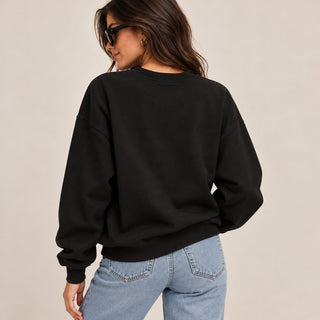 A woman wearing a black crewneck sweatshirt and light blue jeans, photographed from behind against a plain beige studio background.