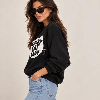 A woman wearing a black crewneck sweatshirt with a white cat silhouette graphic and the text 'Crazy Cat Lady' on the front, paired with light blue jeans and black sunglasses, against a plain light beige background.