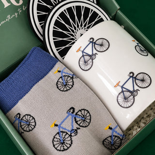 A gift set containing a pair of gray knit socks with a blue and yellow bicycle pattern, a white ceramic mug with a blue bicycle print, and two black-and-white bicycle coasters inside a green box.