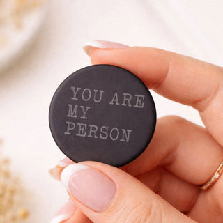 A hand with manicured nails holds a round black button or coaster engraved with the phrase 'YOU ARE MY PERSON' in light gray lettering.