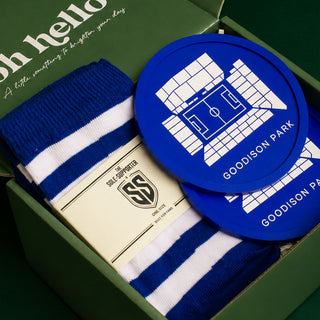 A gift box containing blue and white striped knit socks, a cream-colored self-support card with a shield logo, and two round blue coasters printed with a white stadium graphic and the text GOODISON PARK.