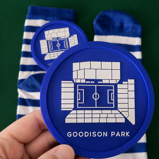 A hand holds a round blue coaster featuring a white graphic of Goodison Park stadium and the text 'GOODISON PARK'. Behind it are blue and white striped socks against a dark green background.