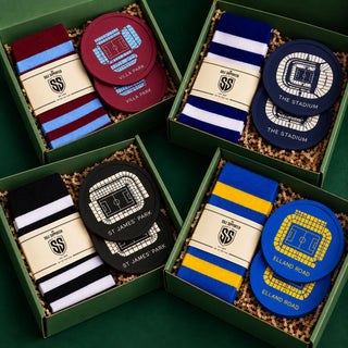 Four green gift boxes arranged on a dark green surface, each containing a pair of striped socks and a circular stadium-themed coaster. The boxes feature designs for Villa Park, The Stadium, St James' Park, and Elland Road