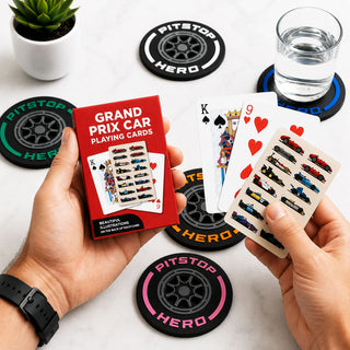 A person holds a red box of Pitstop Hero Grand Prix Car Playing Cards and shows several playing cards with printed race cars. The scene is set on a light marble surface with several black tire-style coasters and a glass of water in the background.