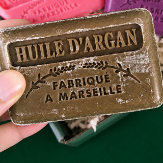 A hand holds a rectangular bar of argan oil soap with embossed French text reading 'HUILE D'ARGAN' and 'FABRIQUÉ A MARSEILLE'. The soap has a distressed bronze-brown finish with white chalky residue.