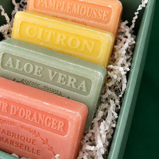Four rectangular artisanal soap bars in different colors, labeled Pamplemousse, Citron, Aloe Vera, and Orange Zing, arranged in a green gift box with white paper shred.