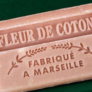 Close-up of two pale pink soap bars with embossed French text reading 'FLEUR DE COTON' and 'FABRIQUE A MARSEILLE' on a dark green background.