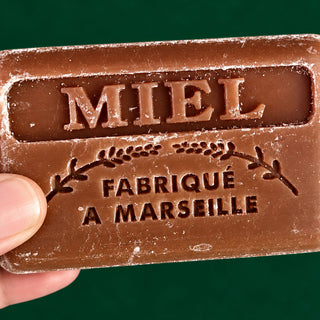 A close-up of a hand holding a rectangular brown bar of soap or wax with embossed text reading 'MIEL FABRIQUÉ A MARSEILLE' against a dark green background.