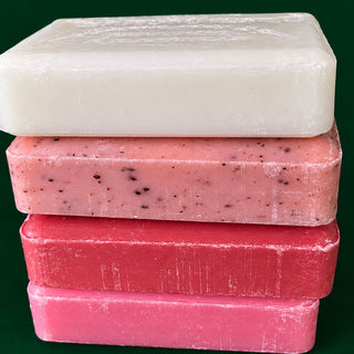 Four stacked rectangular bars of soap in white, pale pink, medium pink, and bright pink, with dark specks visible in some bars, set against a solid dark green background.
