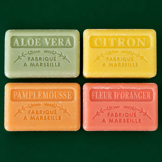 Four rectangular bars of Marseille soap arranged in a 2x2 grid on a dark green background. The soaps are labeled ALOE VERA (light green), CITRON (yellow), PAMPLEMOUSSE (orange), and FLEUR D'ORANGE (pink), each with 'FABRIQUE A MARSEILLE' embossed in the center.