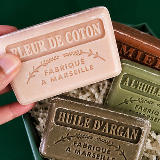 A hand holds a rectangular bar of pale peach soap labeled 'FLEUR DE COTON' and 'FABRIQUÉ A MARSEILLE' in front of three other branded soap bars in a green box with shredded paper filler.