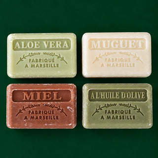 Four rectangular bars of Marseille soap arranged in a 2x2 grid on a dark green background. The soaps are labeled 'ALOE VERA', 'MUGuet', 'MIEL', and 'AL'HUILE D'OLIVE', with 'FABRIQUÉ A MARSEILLE' printed below each.
