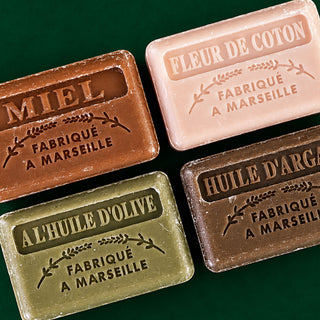 Four rectangular bars of Marseille soap arranged in a 2x2 grid on a dark green background. The soaps are labeled in French with their respective scents: MIEL (beige), FLEUR DE COTON (pink), A L'HUILE D'OLIVE (olive green), and HUILE D'ARGAN (dark brown).