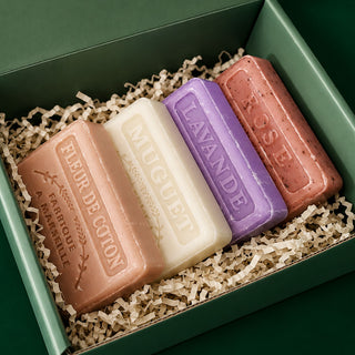Four artisanal bar soaps in different colors (peach, cream, purple, and terracotta) arranged in a green gift box with shredded paper filler. The soaps are embossed with names including 'FLEUR DE COTON', 'MUGUET', 'LAVANDE', and 'ROSÉ'.