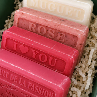 Four decorative soap bars arranged in shredded paper filler. The soaps are embossed with names and phrases including 'MUGuet', 'ROSE', 'I <heart> YOU', and 'UIT DE LA PASSION'.