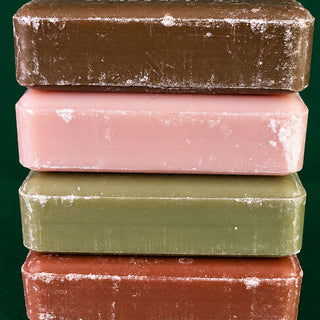 Four rectangular bars of soap stacked vertically, showing different colors: dark brown, light pink, olive green, and terracotta red. Each bar has a slightly rough, textured surface with a white powder residue.